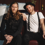 The Lumineers sitting together wearing a suit and a hat and braces 